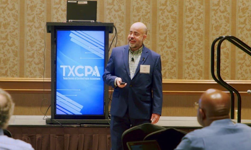 A man in a suit speaks at a podium with a screen displaying "TXCPA," ideal for Instructional Video Production in Memorial City
