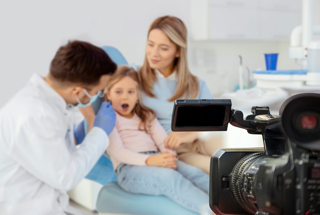 A dentist examining a young girl's mouth while her mother watches. The scene is being captured by a video camera for promotional content.