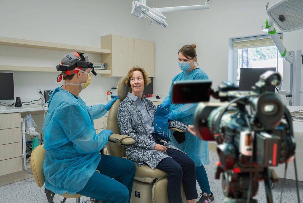 A dental surgeon and assistant in scrubs attending to a smiling female patient in a modern surgical setting. A professional camera is in the foreground, suggesting a procedure or consultation is being filmed for promotion.