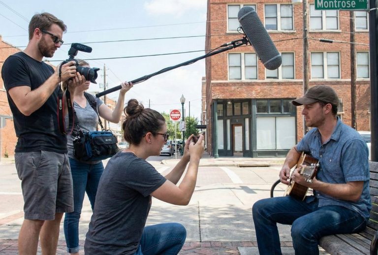 A small film production Houston crew filming a musician playing guitar on a bench in an urban outdoor location.